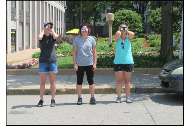 Photo shows 3 women wearing goggles with pin-holes and holding their hands over their ears while standing at the edge of a two-lane street.